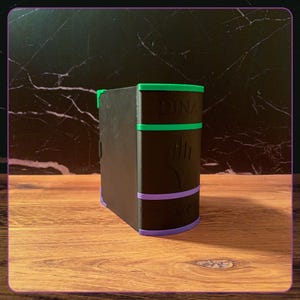 May include: A black rectangular storage box shaped like a book with a green top border and a purple bottom border. The front features the embossed text DINA, a central flame-like symbol, and a row of five circular icons along the lower edge.