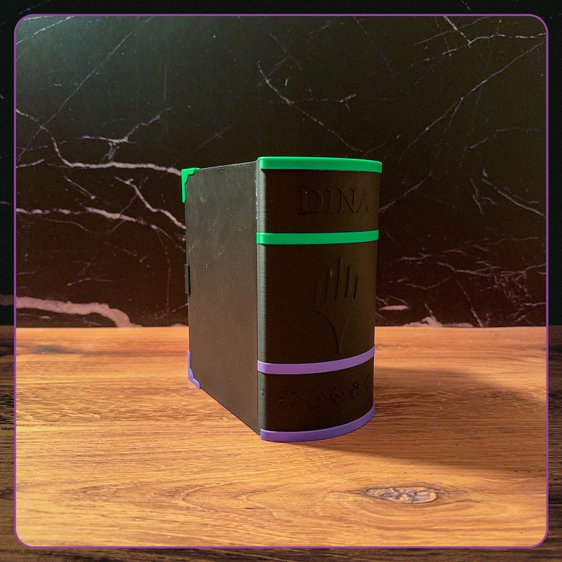 May include: A black rectangular storage box shaped like a book with a green top border and a purple bottom border. The front features the embossed text DINA, a central flame-like symbol, and a row of five circular icons along the lower edge.