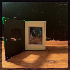 May include: A rectangular display case with a cream-colored frame and a black hinged cover featuring a square cutout. The case holds a single trading card with a dark, illustrated fantasy character and visible text, secured in a clear sleeve.