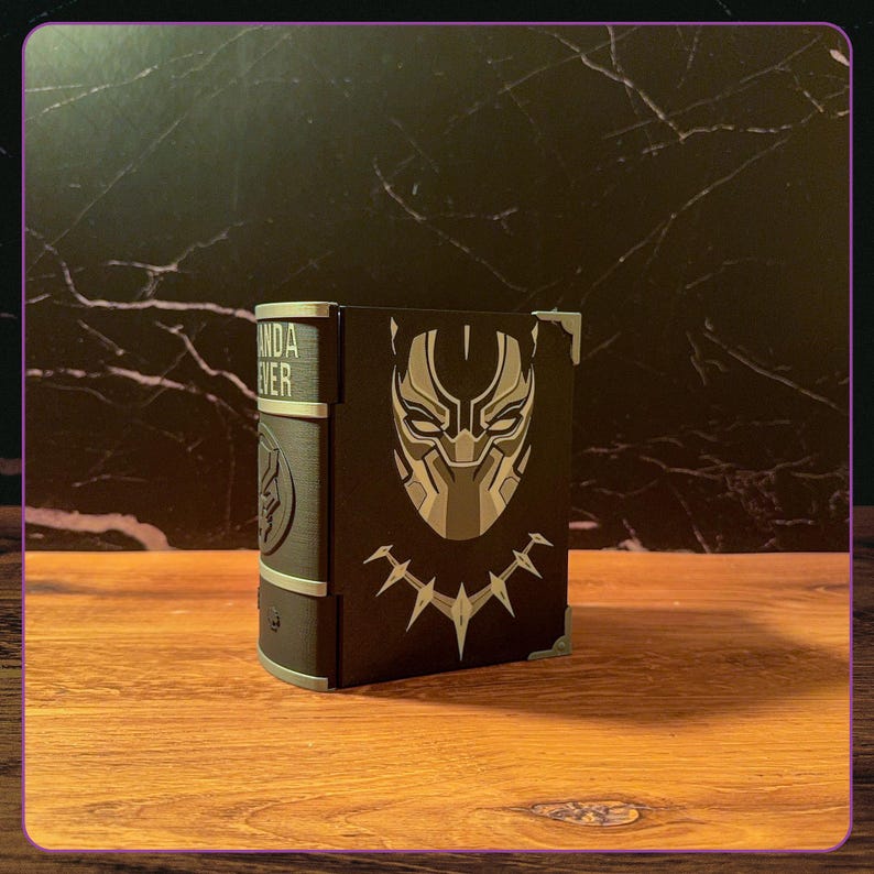 May include: A black and silver decorative book-shaped box featuring a stylized mask graphic and a necklace design on the front cover. The spine displays the text WAKANDA FOREVER in silver lettering, with silver corner accents on the cover edges.