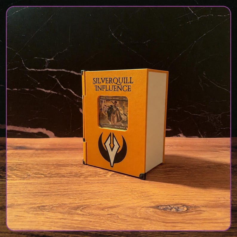 May include: A rectangular orange and white storage box designed to resemble a book, featuring the text "SILVERQUILL INFLUENCE" printed in black above a central window displaying a trading card and a black and white emblem at the bottom.