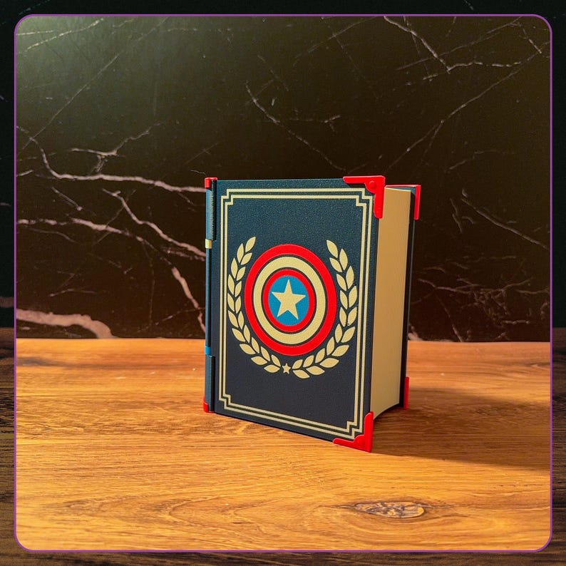 May include: A dark blue rectangular storage box designed to resemble a book, featuring a central circular shield graphic with a white star, red and blue rings, and gold laurel leaves. The box has bright red corner protectors and a blue spine.