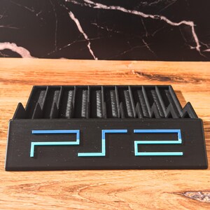 May include: A black PS2 game holder with blue detailing. The holder has multiple slots for game discs and features the letters "PS2" in a stylized font. The holder is made of a matte material and sits on a wooden surface.
