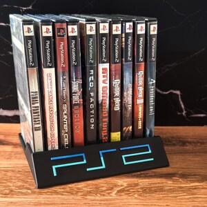 May include: A black PS2 game holder with a blue neon PS2 logo, holding several PlayStation 2 game cases. The game cases have various titles, including "Final Fantasy XI" and "Guitar Hero III."