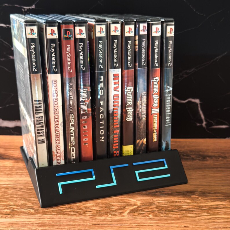 May include: A black metal game stand displaying PlayStation 2 video game cases. The stand has a blue illuminated PlayStation 2 logo. Game titles visible include "Final Fantasy XI", "Guitar Hero III", and "Resident Evil".
