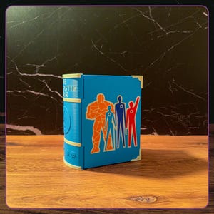 May include: A blue book-shaped storage box featuring the Fantastic Four logo and character silhouettes in orange, blue, and red. The spine displays the text The Fantastic Four with cream-colored corner accents and a textured finish on the surface.