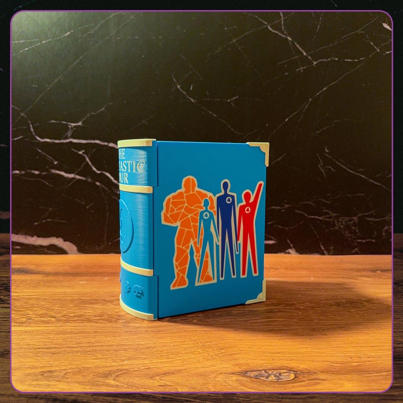 May include: A blue book-shaped storage box featuring the Fantastic Four logo and character silhouettes in orange, blue, and red. The spine displays the text The Fantastic Four with cream-colored corner accents and a textured finish on the surface.