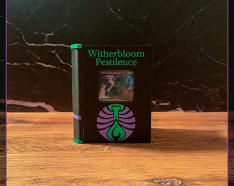 May include: A black rectangular deck box with a green spine and purple accents. The front features the text Witherbloom Pestilence in green, a circular purple and green emblem, and a central window displaying a holographic trading card.