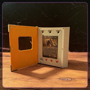 May include: A white and orange plastic display case for a trading card, featuring a hinged cover with a rectangular cutout. The interior holds a single card with the text "TAX" printed below it, alongside three small red heart-shaped icons.