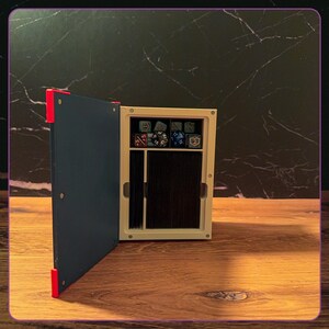 May include: A rectangular storage box with a dark blue hinged cover and bright red corner accents. The interior is off-white, featuring a top compartment filled with various multi-sided gaming dice and a larger lower section holding a deck of cards.