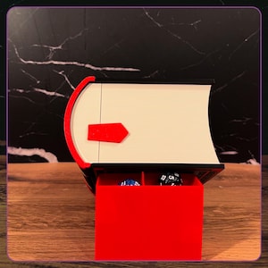 May include: A black and white book-shaped storage box with a red spine and a red latch. The box is open, revealing a red interior compartment divided into two sections, each containing a multi-sided gaming die in blue and black colors.