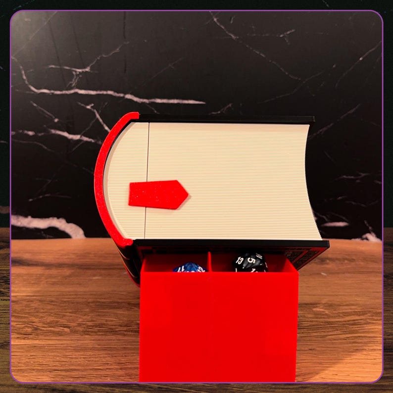 May include: A black and white book-shaped storage box with a red spine and a red latch. The box is open, revealing a red interior compartment divided into two sections, each containing a multi-sided gaming die in blue and black colors.