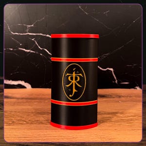 May include: A cylindrical black container featuring a red rim at the top and bottom, with two additional red horizontal bands encircling the middle. A gold oval emblem is centered on the front, displaying the stylized JR Tolkien monogram logo.