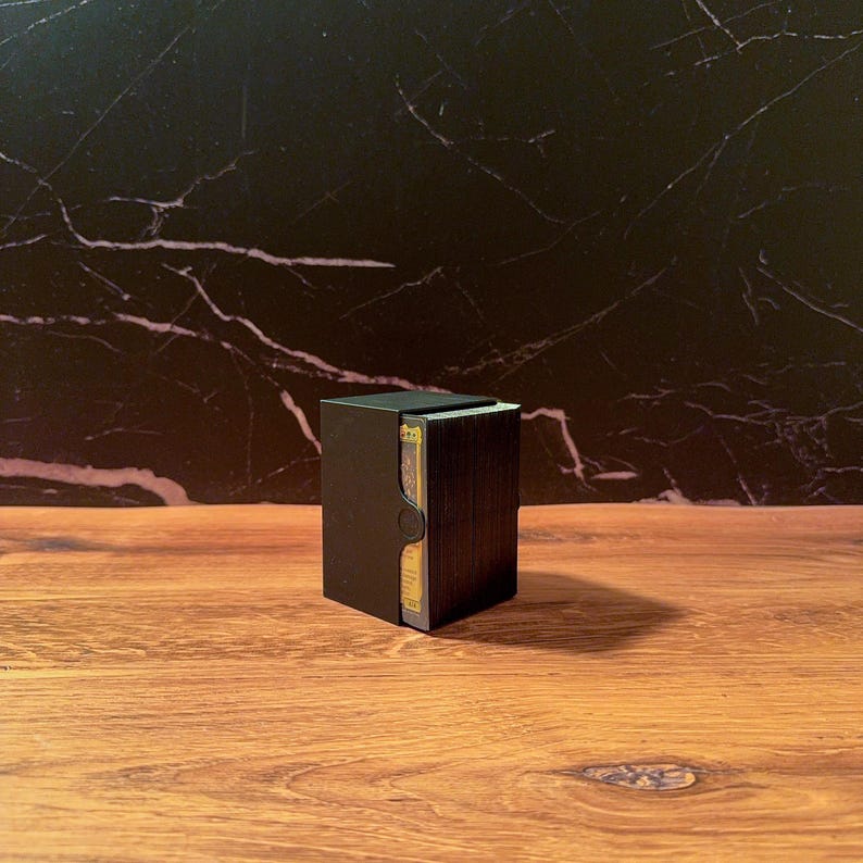 May include: A black rectangular deck box containing a stack of playing cards. The box features a cutout on the front side, revealing the edges of the cards inside. The exterior of the box is solid matte black, providing a sleek and simple storage solution.