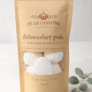 May include: A brown paper bag with a clear window displaying white dishwasher pods. The bag has the text "HEARTHSTONE - HANDMADE" and "dishwasher pods - homemade & non-toxic". Ingredients listed include baking soda, washing soda, citric acid, and salt.