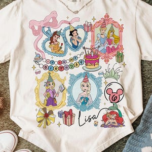 May include: White t-shirt featuring a colorful graphic of Disney princesses in ornate frames, a birthday cake, and the text "It's My Birthday". The name "Lisa" is written in cursive below the images. The shirt has a crew neck and short sleeves.