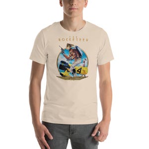 May include: A cream-coloured t-shirt with a graphic of a retro-style rocket pilot and the text "The Rocketeer" in gold. The pilot is in a circular design with a yellow aircraft. The t-shirt has short sleeves.
