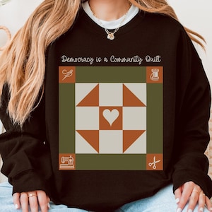 May include: Black sweatshirt with a quilt block design, a heart, triangles, and the text "Democracy is a Community Quilt." The design includes sewing tools and a thread spool. The sweatshirt is a casual, comfortable style.
