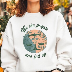 May include: White sweatshirt with the text "We the people are fed up" in a dark green script. The graphic features a balance scale inside a circle divided into two colours, teal and peach, with green leaves around it.