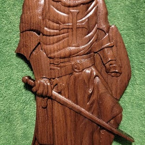 May include: A detailed wooden carving of a knight in full armor, holding a sword and shield. The knight stands before a cross, all crafted from dark brown wood. The carving is set against a green background.