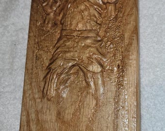 May include: A wooden plaque featuring a carved relief of a figure with outstretched arms. The figure is encased in a block, with details like clothing and facial features visible. The wood has a warm, natural tone.