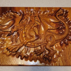 May include: A detailed, hand-carved wooden plaque featuring a dragon. The dragon is depicted with wings, claws, and scales, set against a textured background. The wood has a warm, natural tone, with visible grain and intricate carving details.