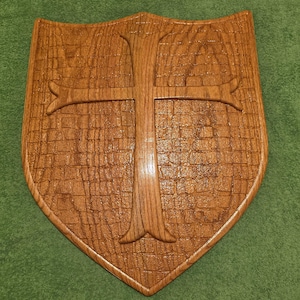 May include: A shield-shaped wooden wall plaque featuring a raised cross in the center. The surface of the plaque has a textured, grid-like pattern carved into the wood, finished with a glossy, warm brown stain that highlights the grain.