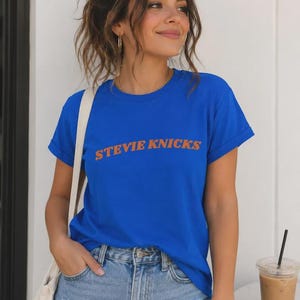 Op de afbeelding: Een felblauw T-shirt met korte mouwen en de tekst STEVIE NICKS in het oranje op de borst gedrukt. Het shirt heeft een ronde hals en opgerolde mouwen, gecombineerd met een lichtblauwe spijkerbroek met versleten details op de bovenbenen.