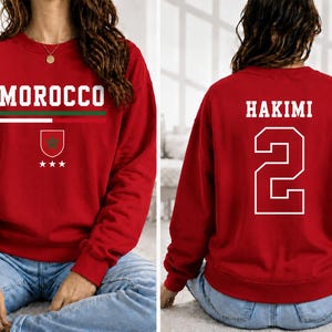 Könnte beinhalten: Ein rotes Sweatshirt mit Rundhalsausschnitt, das auf der Vorderseite den Schriftzug MOROCCO in weißem Text, einen grünen und weißen horizontalen Streifen, ein Wappen mit einem grünen Stern und drei weiße Sterne zeigt. Hinten stehen HAKIMI und die 2.