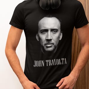 Op de afbeelding: Zwart T-shirt met een zwart-wit afbeelding van een mannengezicht en de tekst "JOHN TRAVOLTA" eronder. De persoon draagt een zwarte koptelefoon. Het T-shirt heeft een ronde hals.