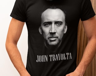 May include: Black t-shirt featuring a black and white image of a man's face, with the text "JOHN TRAVOLTA" printed below the image in a light grey font. The person is wearing black headphones.