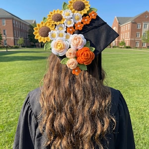 May include: A black graduation cap adorned with a vibrant, handmade crochet floral arrangement. The flowers include yellow sunflowers, white daisies, and orange and peach roses. The cap is worn by a person in a black graduation gown.