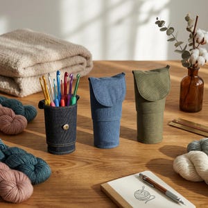 May include: Three cylindrical fabric cases in dark blue, medium blue, and olive green, each with a fold-over flap. The dark blue case holds a collection of colorful metal crochet hooks. The cases feature a structured design with a metal snap closure.