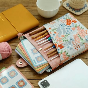 May include: A fabric pouch with a floral embroidery pattern containing several crochet hooks and a stack of colorful instructional cards. The pouch features a snap closure and is surrounded by a yellow notebook, a white mug, yarn, and a tablet.
