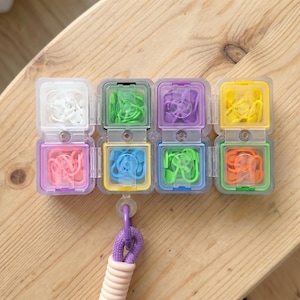 May include: A set of eight small, square, clear plastic storage containers arranged in two rows of four, each filled with colourful plastic stitch markers in white, pink, green, blue, purple, yellow, and orange, attached to a purple cord.