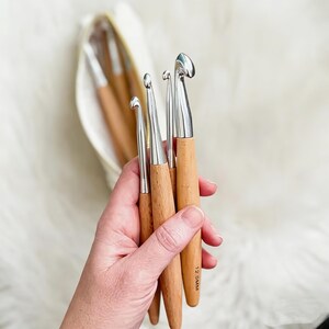 Puede incluir: Un juego de cuatro ganchillos con cabezas de metal plateado pulido y mangos de madera de color marrón claro liso. El ganchillo más grande tiene la medida 12.0mm impresa en la madera. Los ganchillos se sostienen juntos en un pequeño grupo.