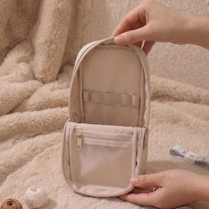 May include: A beige upright organizer case with a zippered opening, featuring an interior elastic band for holding items and a clear zippered pocket. The case is shown open, revealing its internal storage compartments and light-colored lining.
