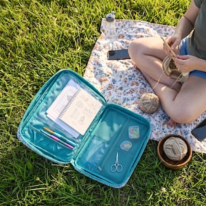 May include: A teal zipped craft organiser case containing a spiral-bound notebook, two crochet hooks, a pair of small silver scissors, and two small clear containers. The case features interior mesh pockets and clear plastic sleeves for storage.