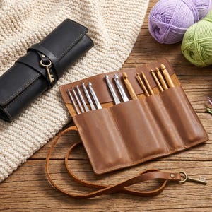 May include: A black leather crochet hook case with a key closure and a brown leather crochet hook roll. The roll is open, displaying various crochet hooks. Several balls of yarn in purple, green, and cream are in the background.