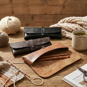 May include: A still-life image of knitting and crochet supplies on a wooden surface. Includes yarn balls in various colors, leather cases with the name "Isabella" embossed, a mug of tea, scissors, and a pattern book.