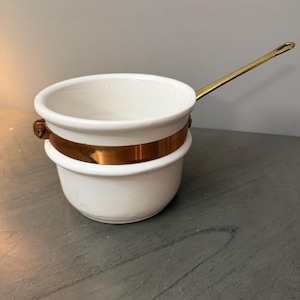 May include: A white ceramic saucepan featuring a polished copper band around the exterior and a long, slender brass handle attached with a metal bolt. The vessel has a rounded base and a wide, open top, showcasing a clean, functional design.