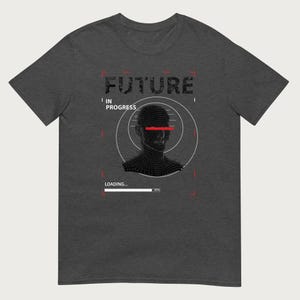 Op de afbeelding: Een donkergrijs t-shirt met een grafische print van een menselijk hoofd in wireframe-stijl. Het ontwerp bevat de tekst FUTURE IN PROGRESS en LOADING... met een voortgangsbalk op 87 procent en een rode lijn over de ogen van het hoofd.
