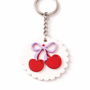 May include: A cherry-themed keychain featuring a white scalloped background with a red cherry design and a purple bow. The keychain has a silver chain and ring.