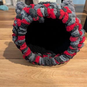 May include: A handmade cat bed in a cave shape. The bed is made of a soft, chunky yarn in a mix of red, gray, and black. The interior is black, and the bed has ear-like protrusions at the top.