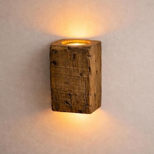 May include: A wooden wall sconce with a square shape. The sconce is made from a weathered, brown wood block, with a recessed top that holds a warm, glowing light. The light casts a soft glow on the wall above and below the sconce.