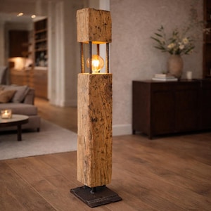 Può includere: Lampada da terra rustica con un palo di legno rettangolare e una parte superiore quadrata. La lampada presenta un apparecchio a gabbia con una lampadina visibile. La base è una piastra di metallo quadrata e scura. Il legno ha un aspetto naturale e invecchiato. Alta circa 150 cm.