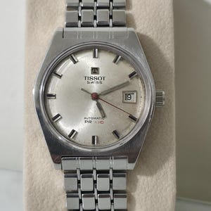 Peut inclure: Une montre-bracelet automatique Tissot argentée avec un bracelet à maillons métalliques. Le cadran est blanc cassé avec des index argentés, un guichet de date à trois heures et l'inscription TISSOT SWISS AUTOMATIC PR 516 imprimée dessus.