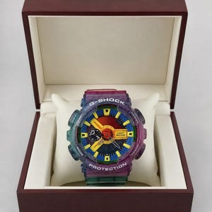 Puede incluir: Un reloj G-Shock multicolor con correa y caja transparentes, presentado en una caja de madera oscura. La esfera del reloj presenta detalles en amarillo, azul y rojo. El reloj se muestra sobre un cojín blanco.