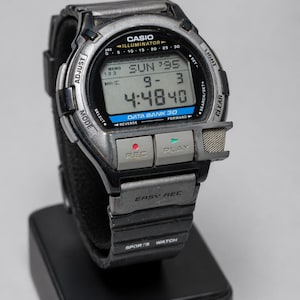 May include: A silver and black Casio Data Bank 30 sports watch. The digital display shows the time, date, and day. The watch has buttons for adjusting, selecting, and recording. The watch is on a black stand.