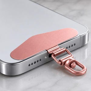 May include: A rose gold adhesive phone tether tab attached to the bottom of a silver smartphone. The accessory features a flat, curved metallic plate that adheres to the device and includes a small, integrated swivel lobster clasp hook.